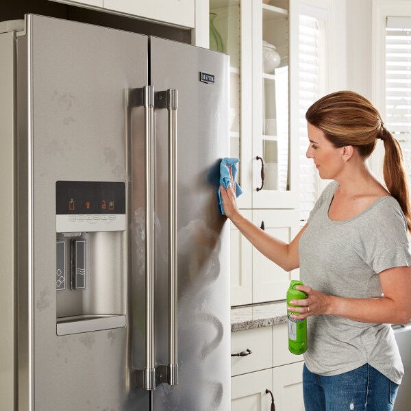 A woman wiping down the outside of a refrigerator door with stainless steel cleaner.