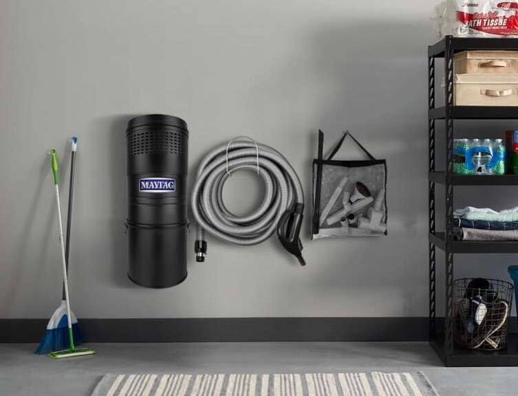 A close up of a Maytag® central vacuum and its accessories on a wall in a garage.
