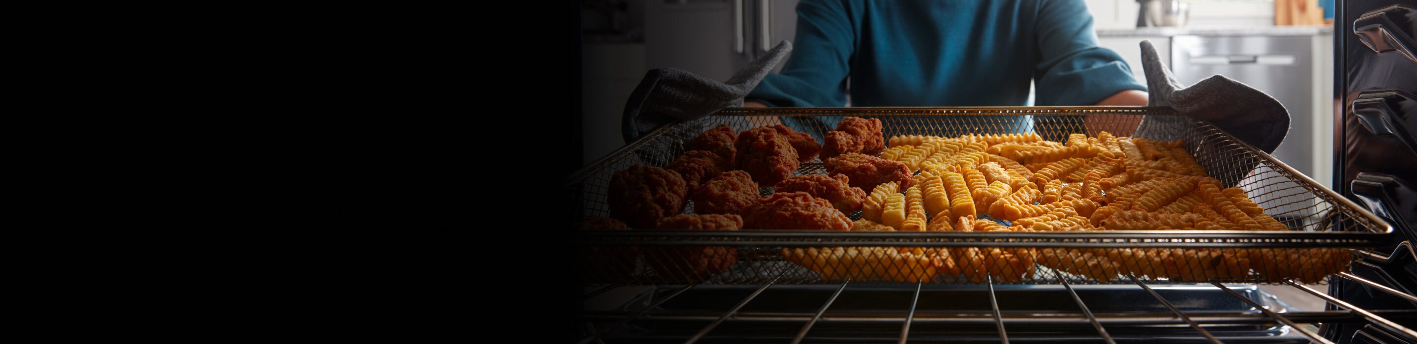 Person pulling an air fryer basket with cooked chicken nuggets and crinkle cut fries out of an oven