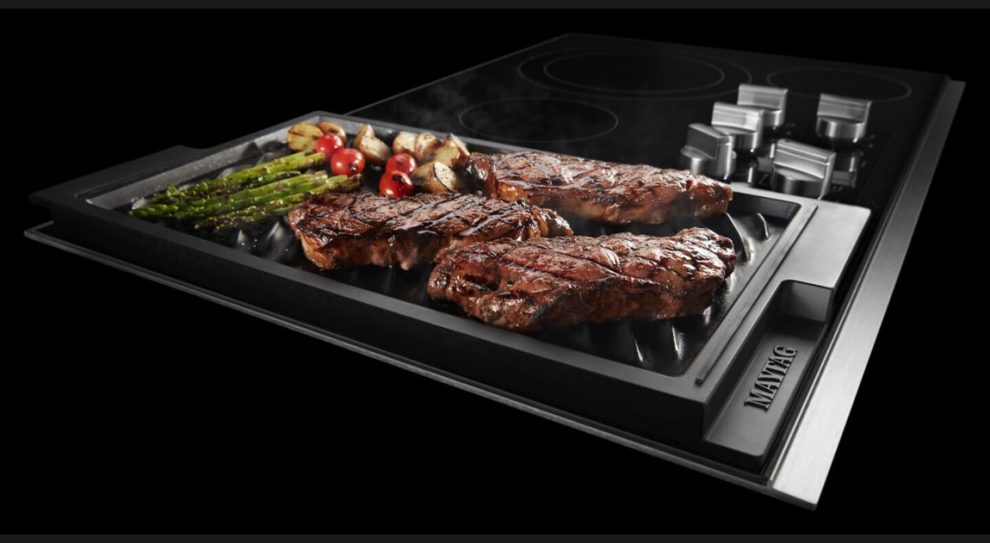 Steaks and vegetables cooking in a cast iron grill pan on a Maytag® electric cooktop