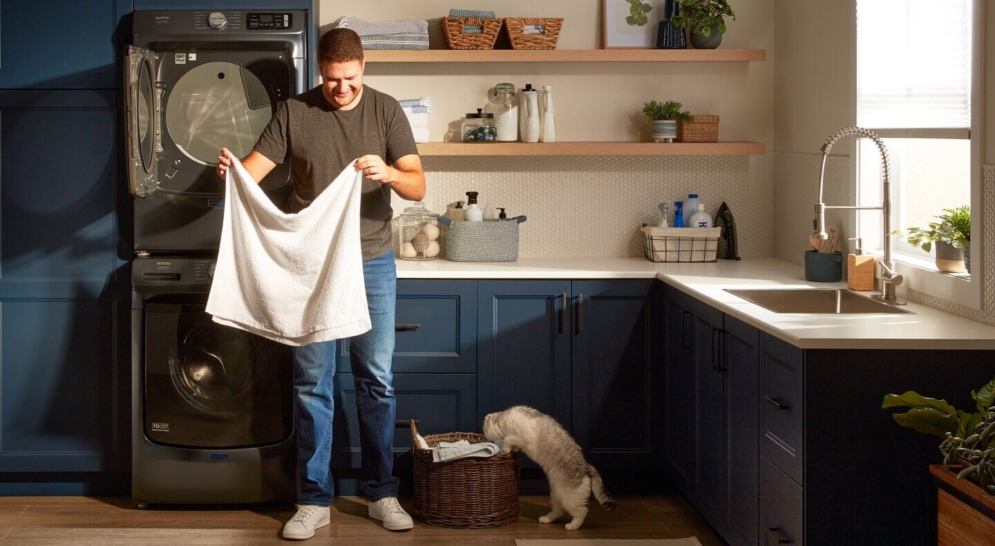 A man folding a white towel in front of his washer and dryer while a cat watches