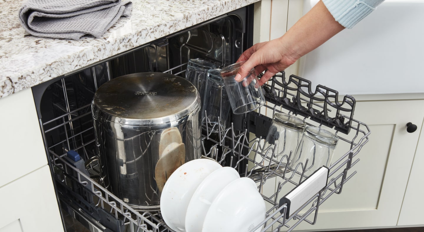 Person loading glass cup into a Maytag brand dishwasher. Person loading glass cup into a Maytag brand dishwasher.