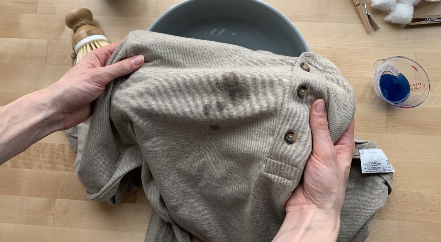 Gray shirt with grease stains and a cup of blue detergent next to it