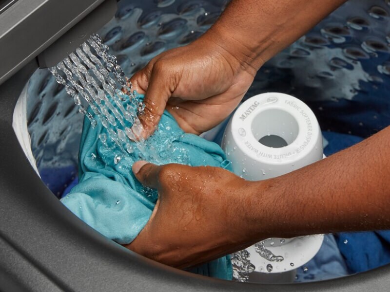 A person adding laundry to a Maytag® top load washing machine filling with water