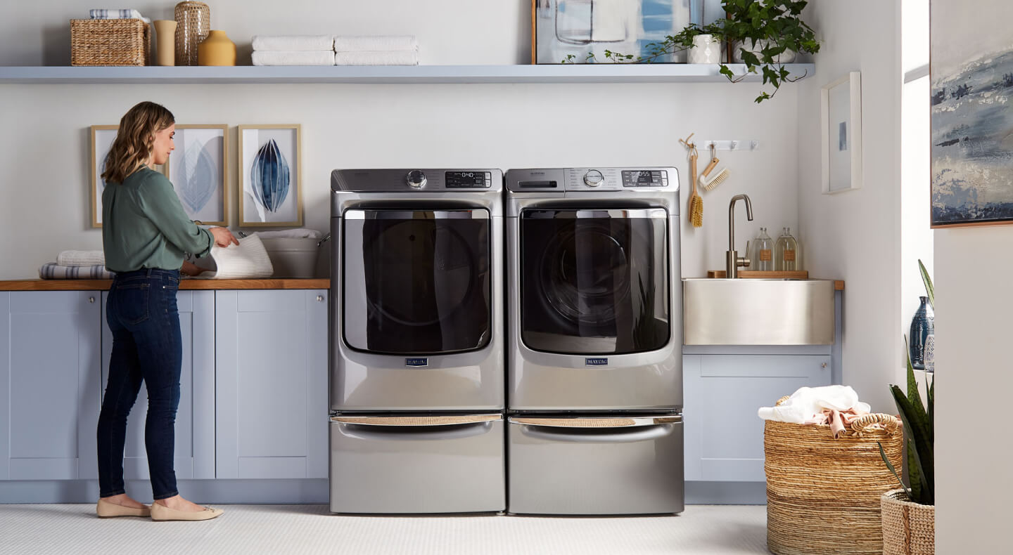 A woman folding laundry next to Maytag® side-by-side washer and dryer set in a modern laundry room A woman folding laundry next to Maytag® side-by-side washer and dryer set in a modern laundry room