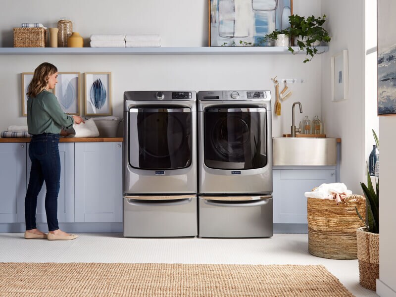 A woman folding laundry next to Maytag® side-by-side washer and dryer set in a modern laundry room A woman folding laundry next to Maytag® side-by-side washer and dryer set in a modern laundry room