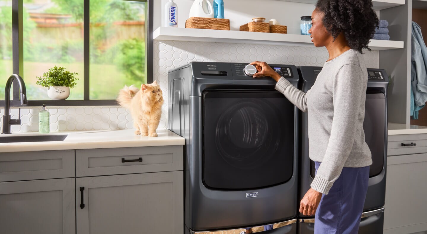 Someone operating the controls of their Maytag® Pet Pro washer as their cat stands nearby