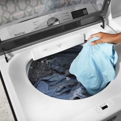 Person adding a blue garment to a white, top-load Maytag® washer