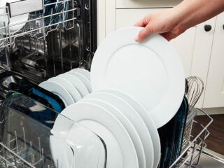 A hand loading a white plate into a dishwasher rack