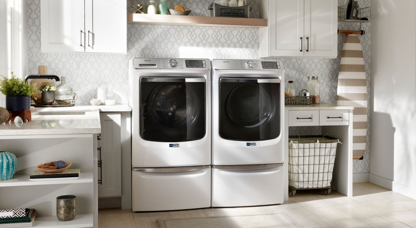 White Maytag® front load washer and dryer pair on pedestals in a laundry room