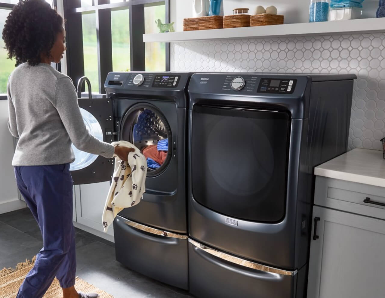 A person putting a blanket with paw prints into a front load washer next to a front load washer, both on pedestals