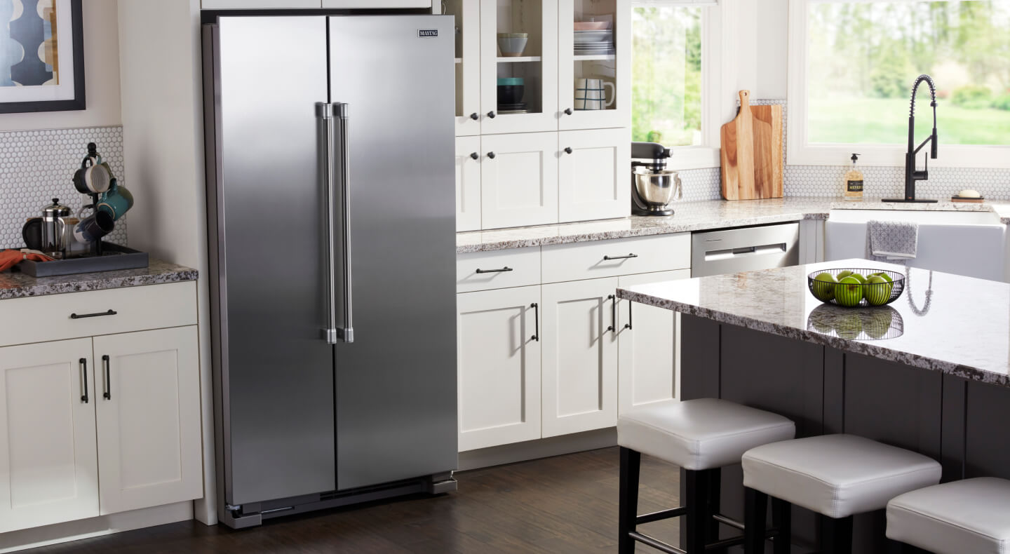 A stainless steel side-by-side refrigerator in a kitchen with white cabinetry and grey granite countertops