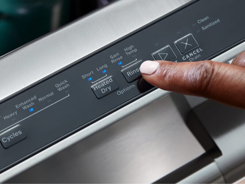 A finger pressing the rinse button of a dishwasher’s control panel