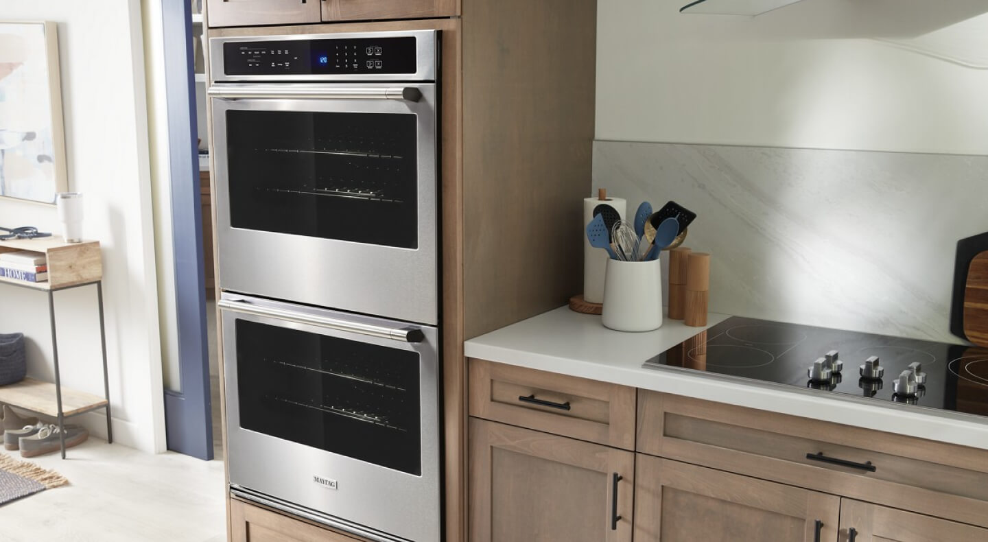 A Maytag® double wall oven installed next to brown cabinets with white countertops and a cooktop with five knobs