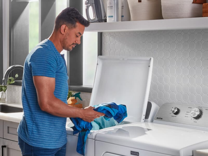 A person adding laundry items to a Maytag® top load washer in a modern laundry room A person adding laundry items to a Maytag® top load washer in a modern laundry room