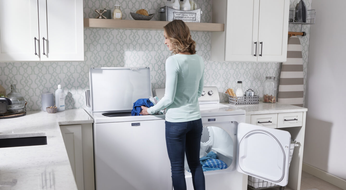 A person adding laundry items to a Maytag® top load washer in modern laundry room