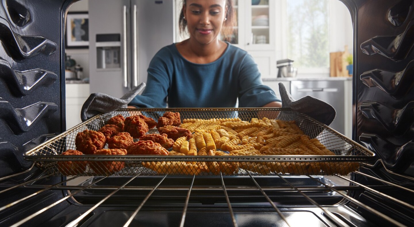 A person pulling an air fry basket filled with fries and chicken wings out of the oven. A person pulling an air fry basket filled with fries and chicken wings out of the oven.