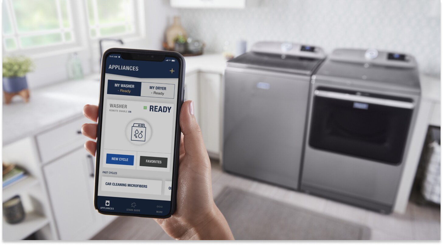 Person using the Maytag™ App standing in front of a gray top-load laundry pair Person using the Maytag™ App standing in front of a gray top-load laundry pair