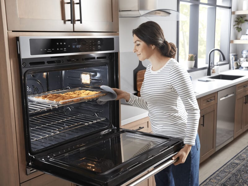 A person taking a sheet pan of food out of a Maytag® single oven in a modern kitchen