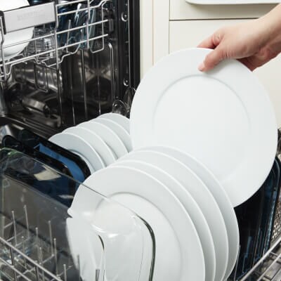 Person placing a white dinner plate in the bottom rack of a dishwasher