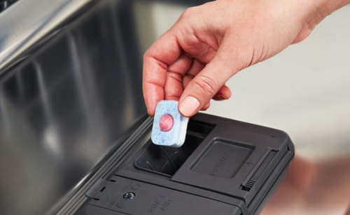 A hand placing a dishwasher pod into a dishwasher dispenser