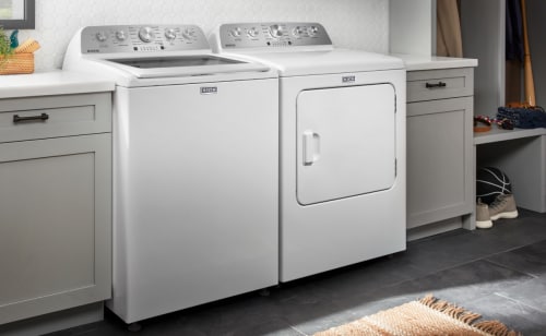 White Maytag® top load washer and front load dryer side by side in a laundry room next to shelves