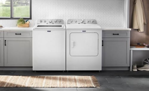 Overview of a laundry room with a top load washer, front load dryer, gray cabinets, and white backsplash tile.