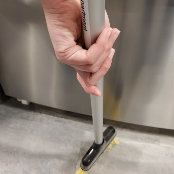 A broom sweeping the floor behind a refrigerator.