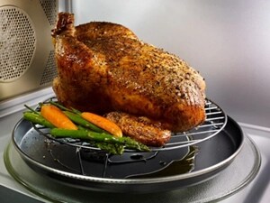 A whole chicken with vegetables cooking on a grilling accessory inside of a microwave