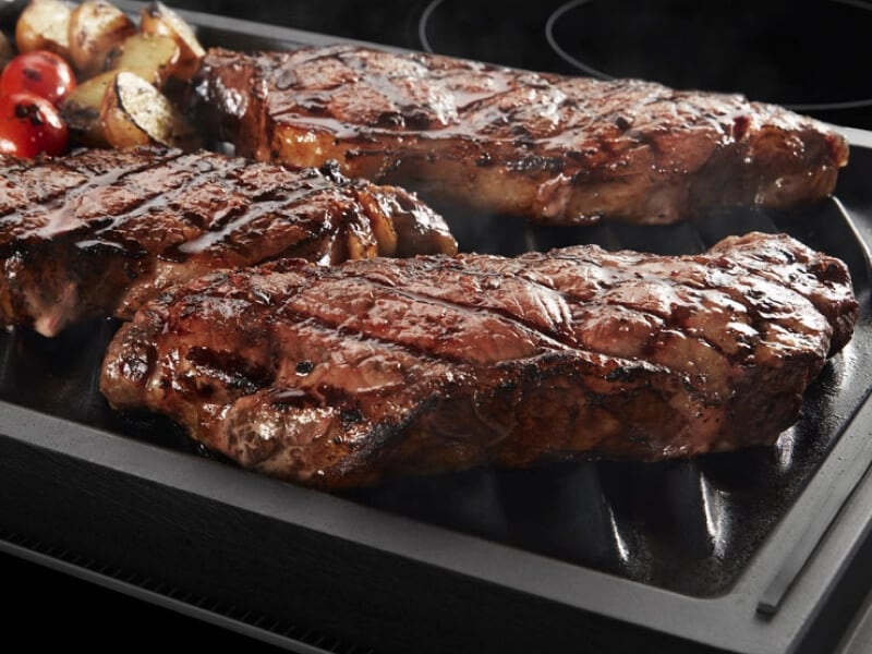 Three grilled steaks and vegetables on a grill pan