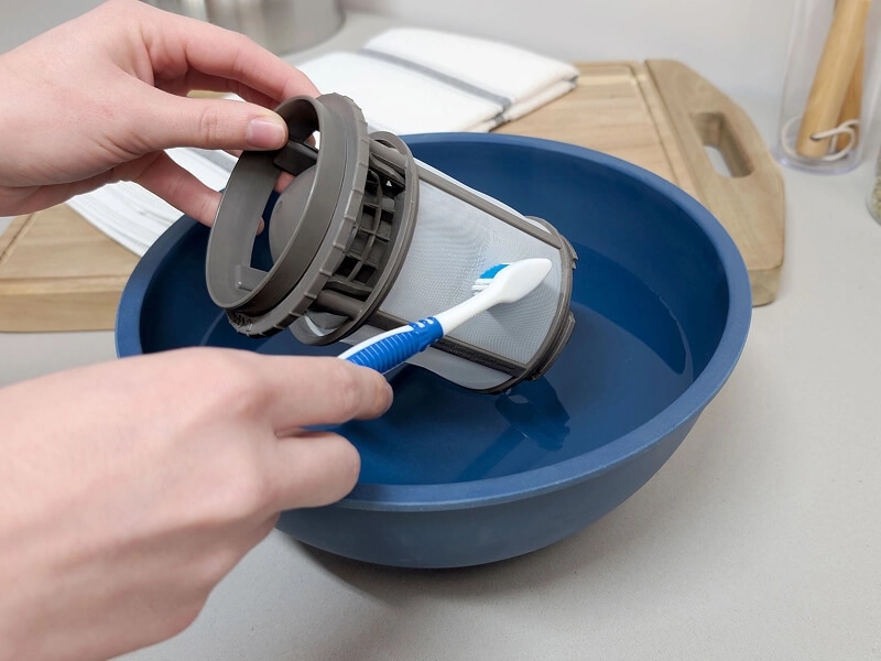 A hand using a toothbrush to scrub a dishwasher filter