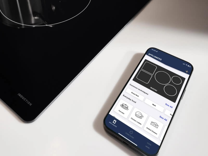 Phone next to a cooktop open to the Maytag® App