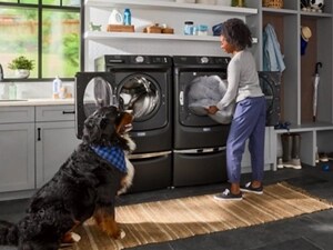 A person unloading a dog bed from dryer on pedestal next to a front load washer on a pedestal with a dog looking on