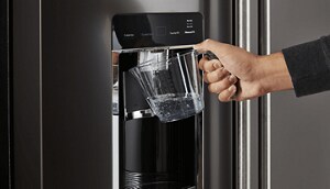 A hand holding a measuring cup under a refrigerator water filter to flush the new system