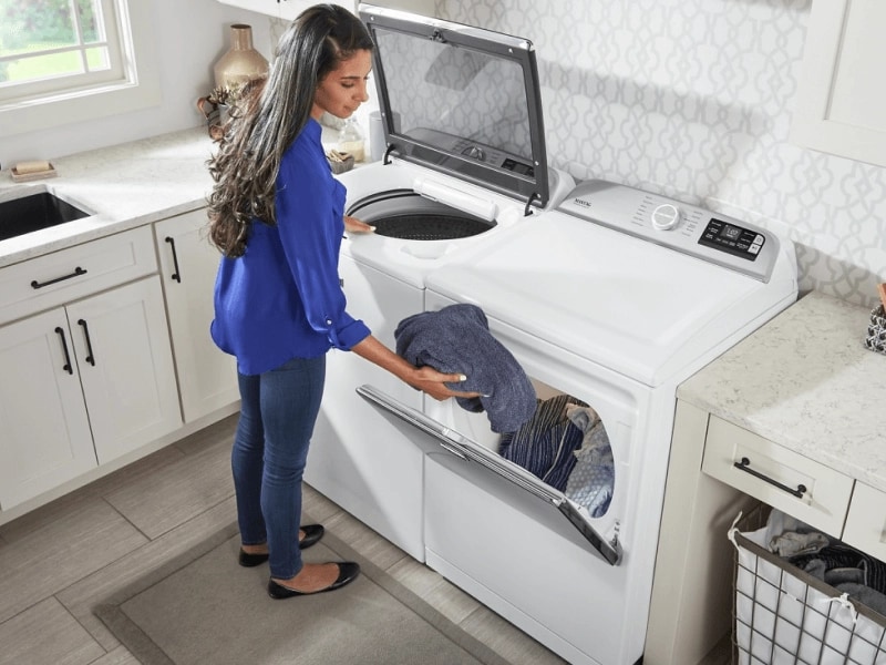 Person in a laundry room with white cabinetry, putting wet laundry in a white Maytag® dryer