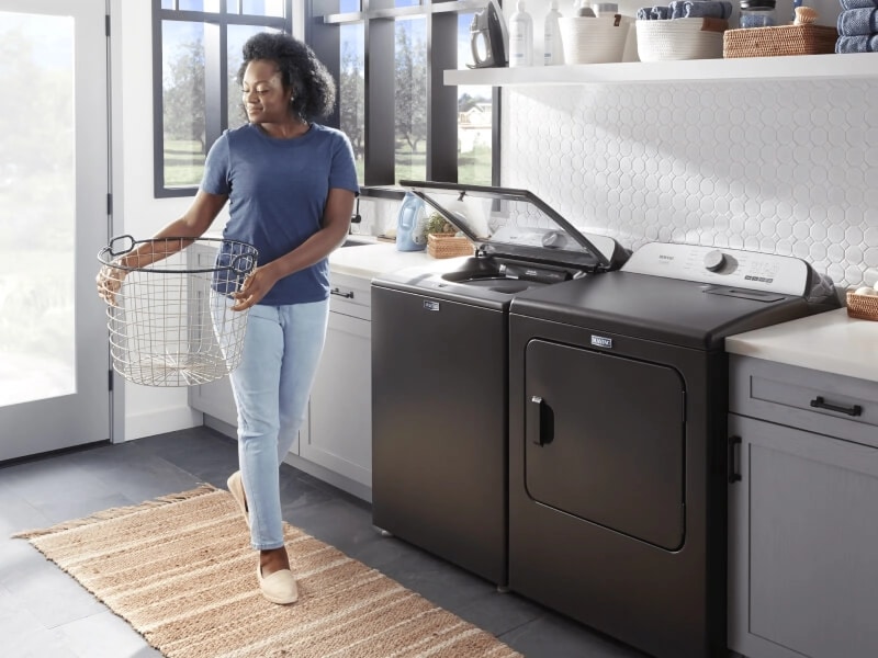 A person holding a laundry basket in a laundry room with a Maytag® top load washer and dryer pair
