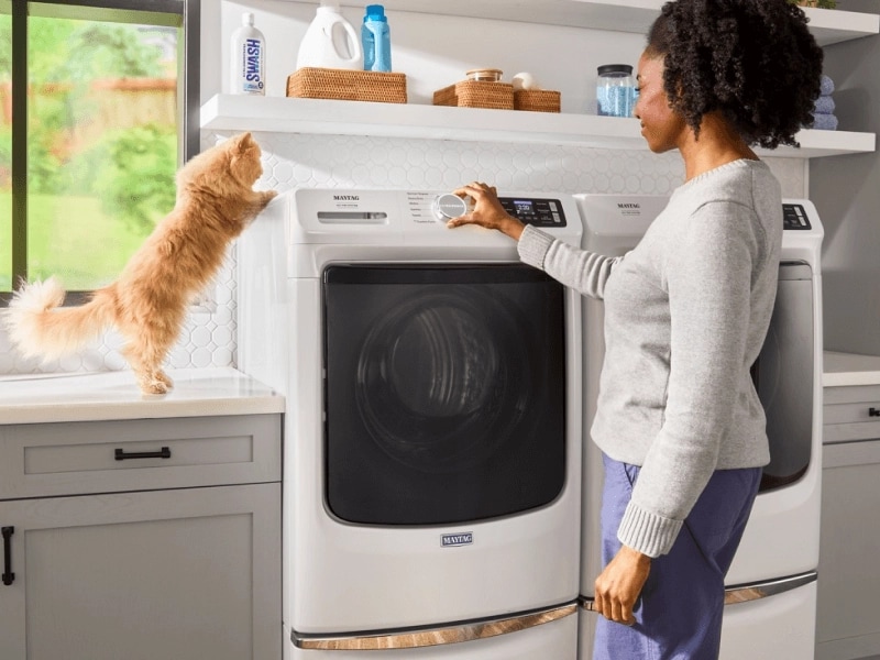 A cat crouching on top of a washing machine as a person adjusts the settings
