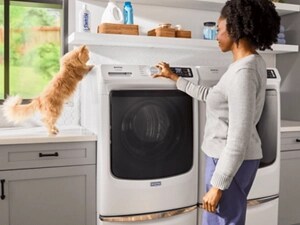 A cat crouching on top of a washing machine as a person adjusts the settings