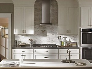 A full view of a modern kitchen with an island in the middle, white cabinetry, a stainless steel and glass wall-mount range hood, a cooktop and dual ovens